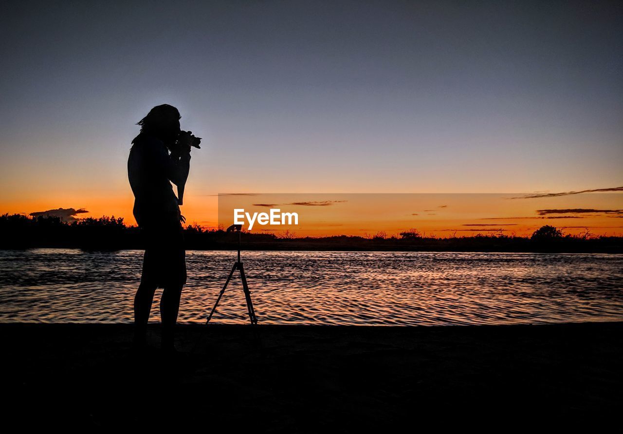 SILHOUETTE MAN PHOTOGRAPHING SEA AGAINST ORANGE SKY