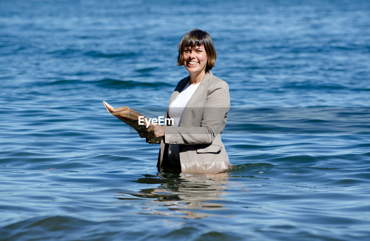 Portrait of smiling young woman holding newspaper while standing in sea