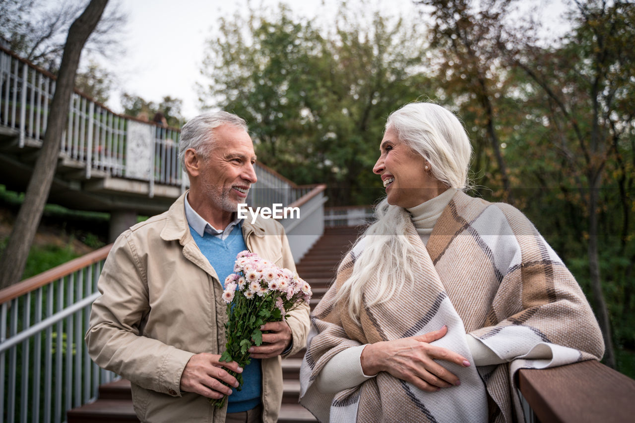 Smiling man holding bouquet standing with woman against trees