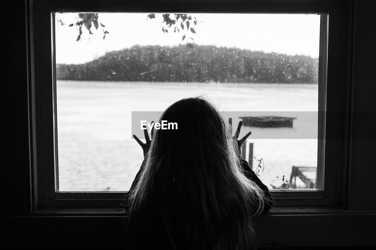 Rear view silhouette of young girl looking through window at a lake