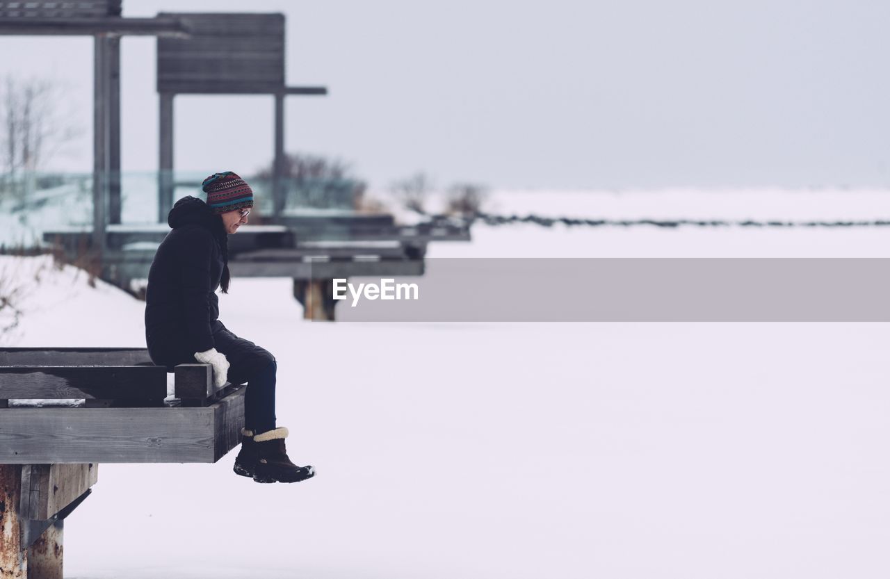 Side view of woman sitting on pier against sky during winter