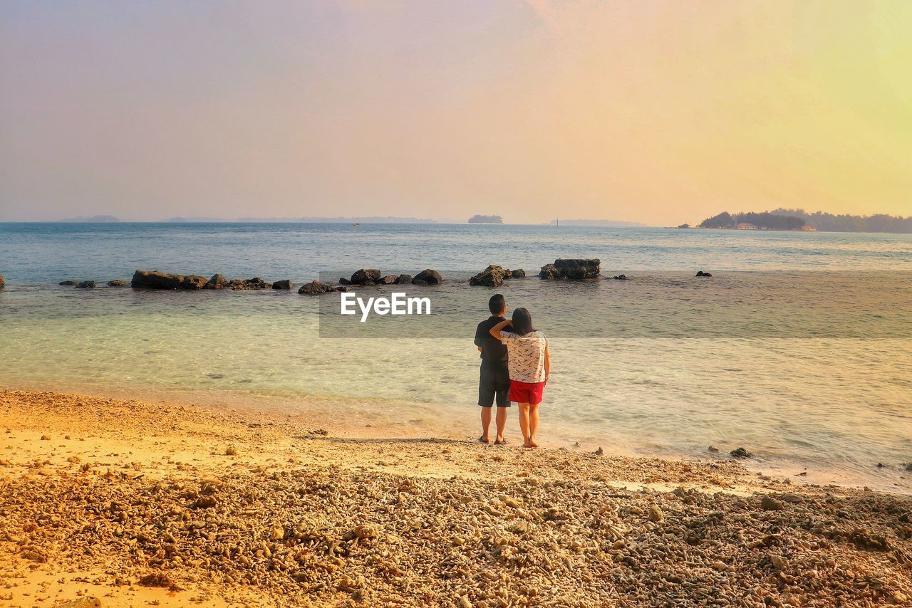 REAR VIEW OF COUPLE STANDING ON BEACH AGAINST SKY