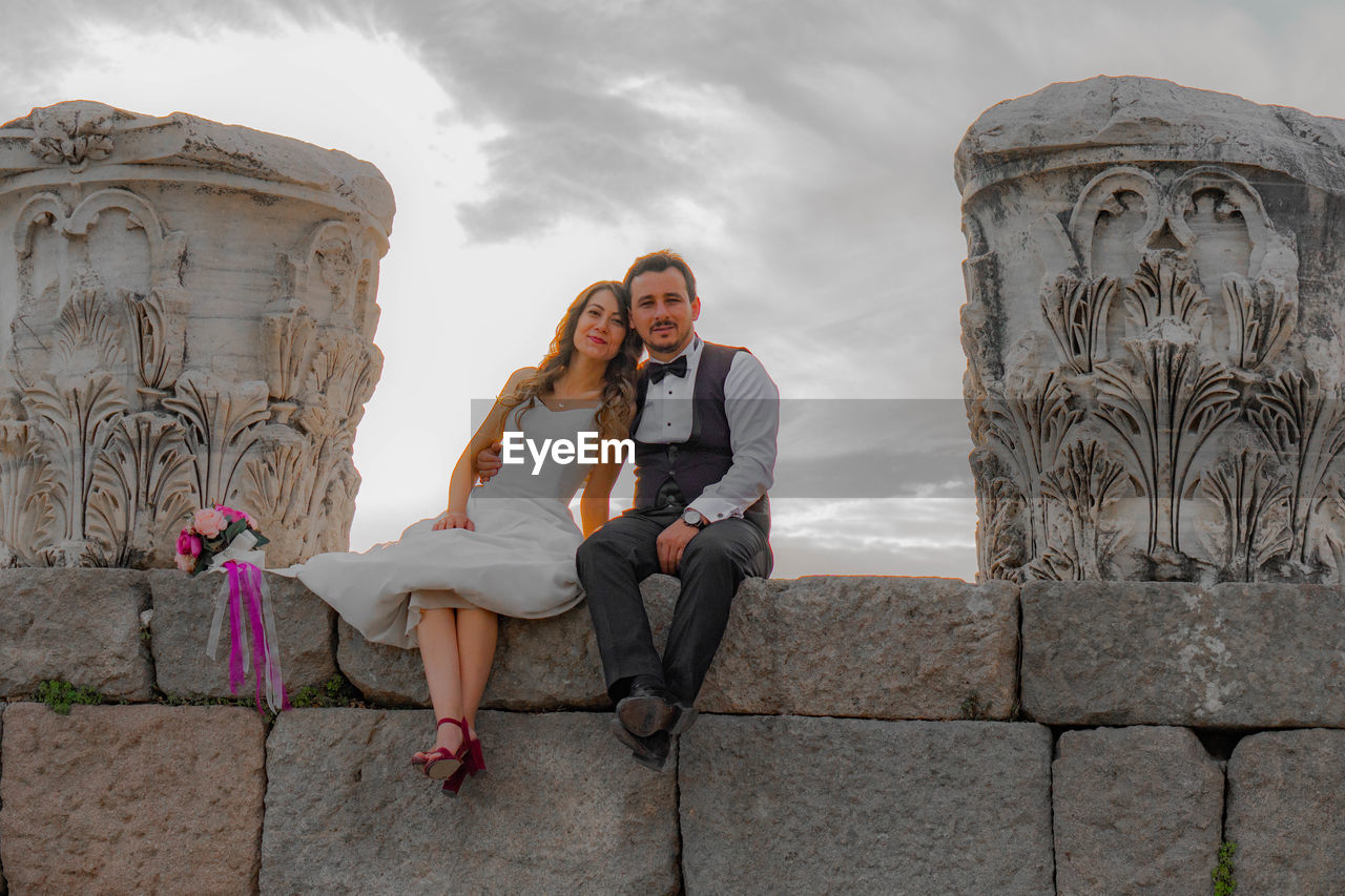 Portrait of smiling bride with groom sitting on stone wall at old ruin