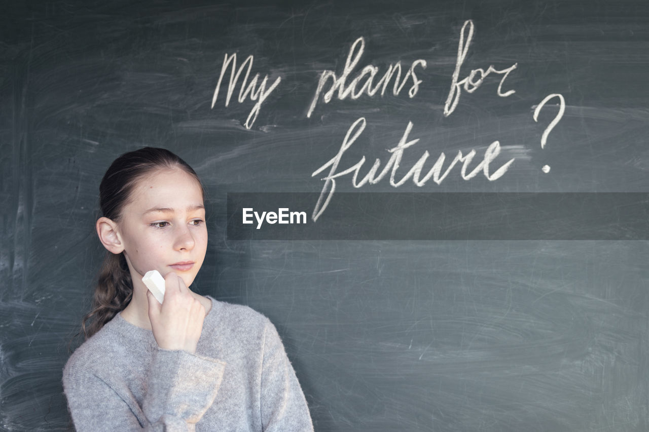 Girl looking away while standing by blackboard with text in classroom
