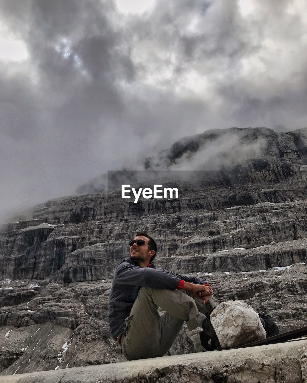 Low angle view of young man sitting on mountain against cloudy sky