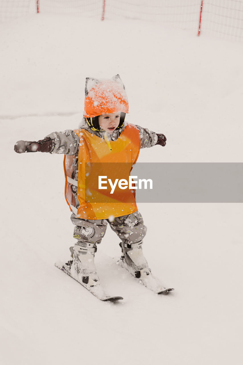 Full body of adorable little child in warm winter overalls and protective helmet standing on skis while practicing skiing on snowy terrain