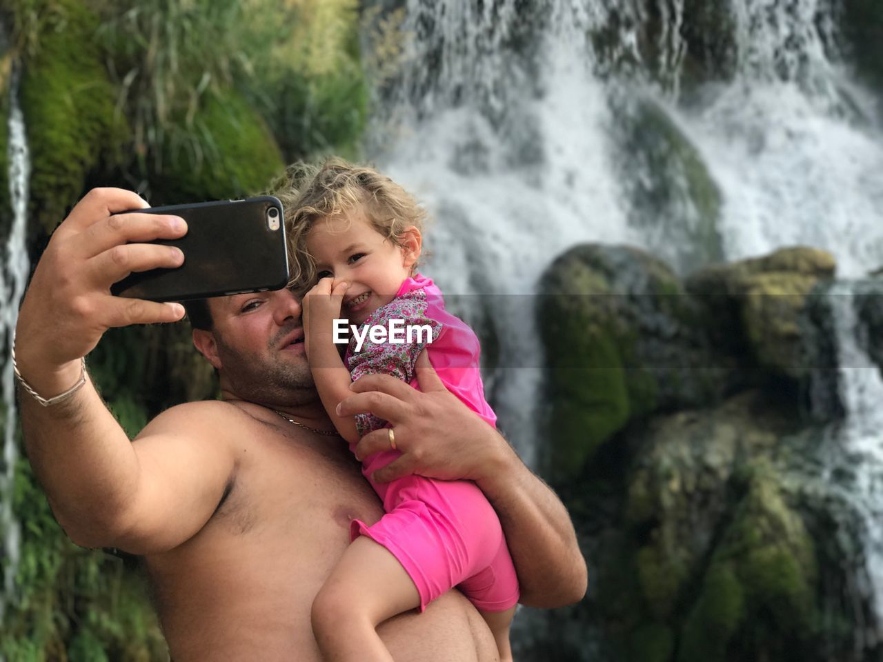 Shirtless father with daughter taking selfie through mobile phone against waterfall