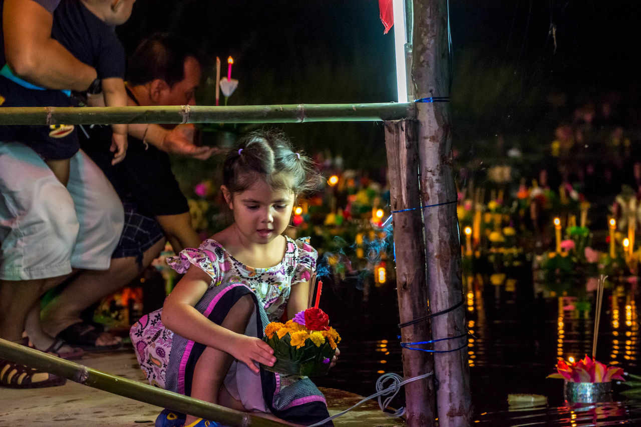 Girl holding flowers while kneeling by lake during loi krathong at night