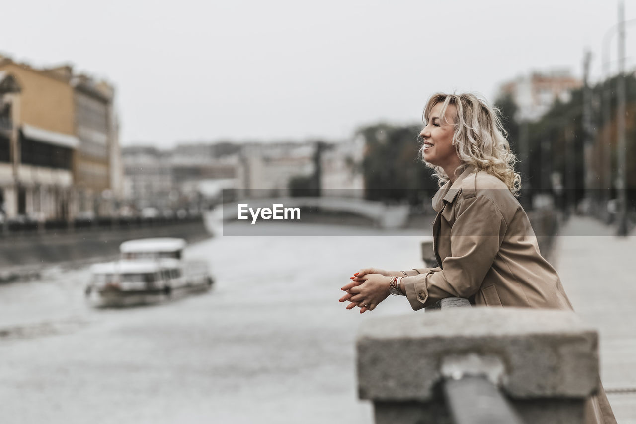 A middle-aged blonde woman with curly hair stands on the river embankment in the city.