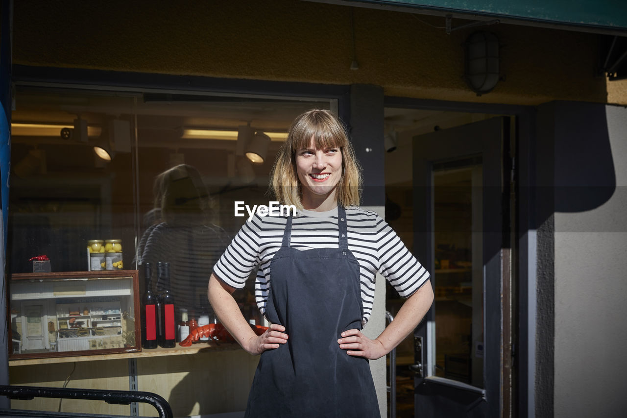 Confident female owner looking away while standing with hands on hips outside store