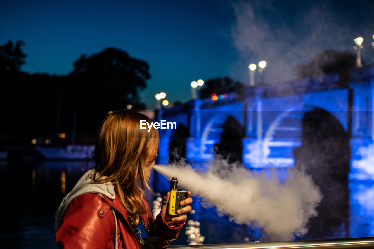 Side view of woman smoking electric cigarette in illuminated lights at night