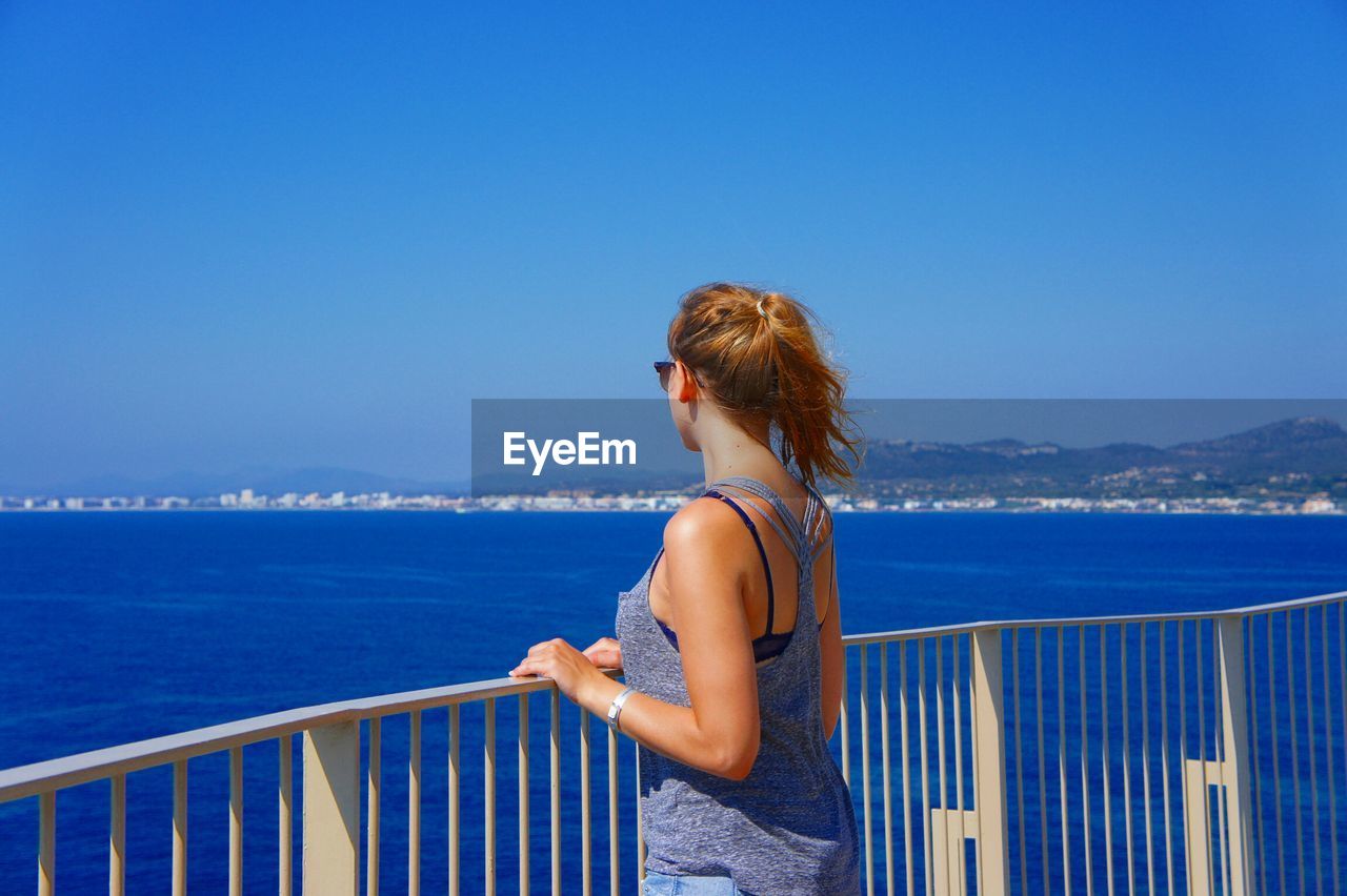 Young woman by railing looking at sea on sunny day