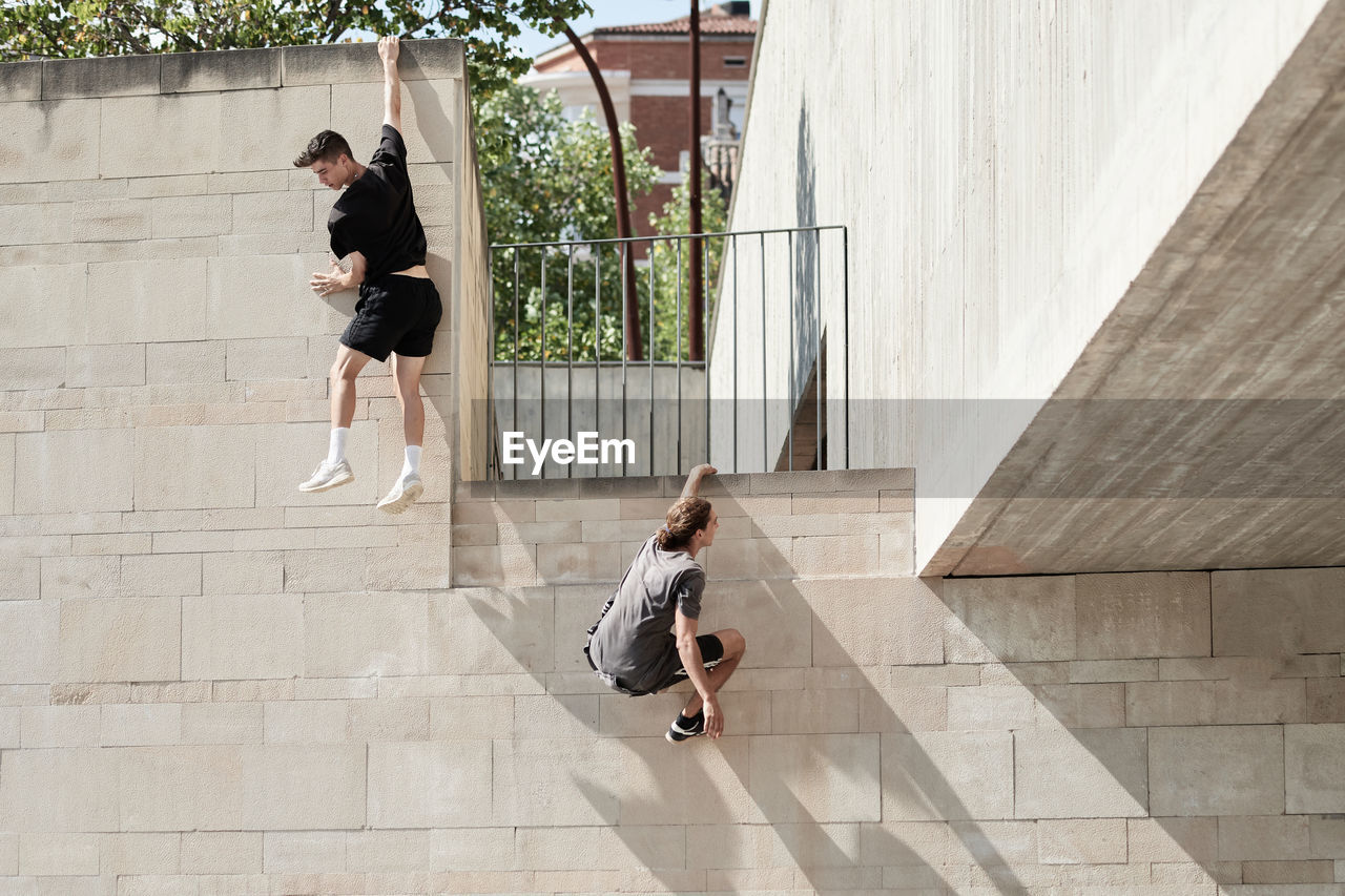 Back view of brave male friends hanging on wall of concrete building while performing risky trick and doing parkour in city