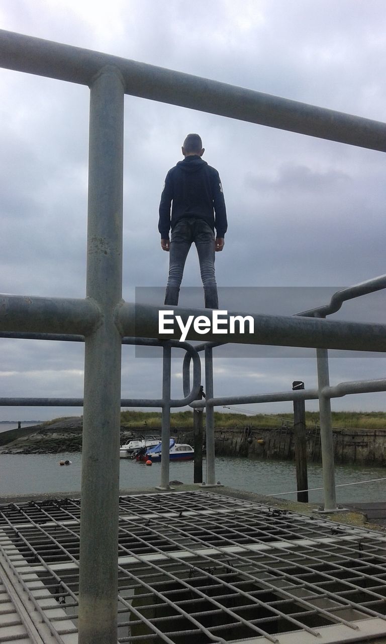 REAR VIEW OF MAN STANDING ON RAILING AGAINST SKY