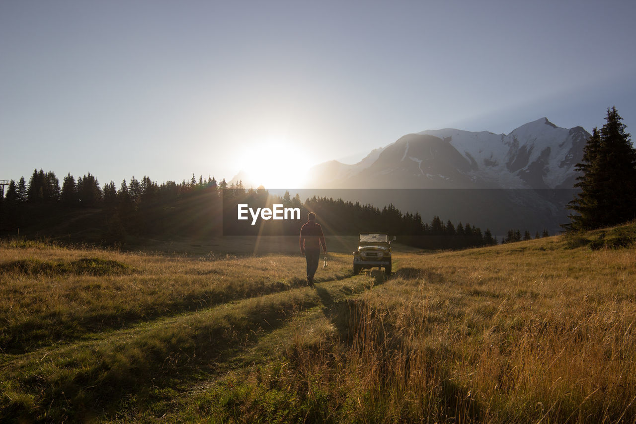 Man walking towards iconic car in a field against mountains during sunrise