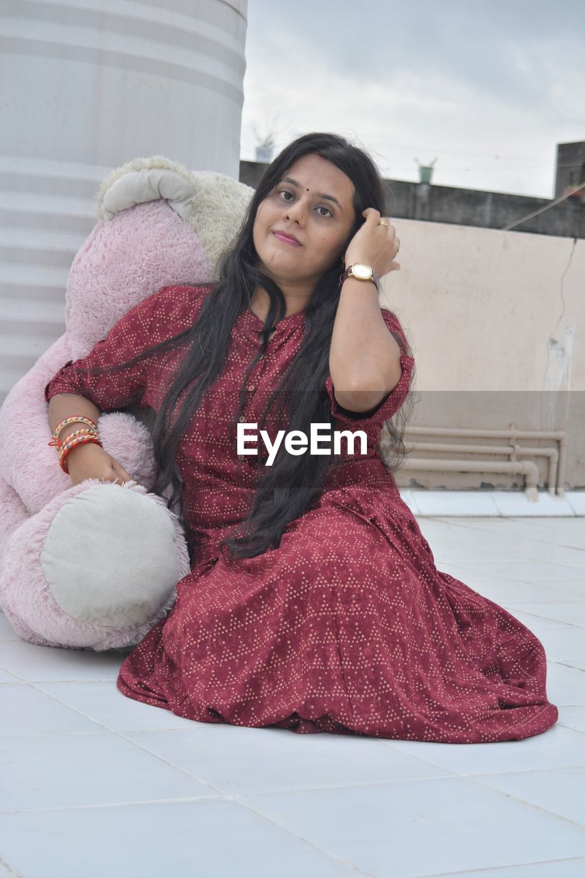 one person, women, pink, adult, young adult, clothing, front view, sitting, long hair, dress, portrait, hairstyle, architecture, lifestyles, full length, textile, looking at camera, fashion, leisure activity, smiling, photo shoot, looking, brown hair, day, built structure, building exterior, red, relaxation, outdoors, person, emotion, female, casual clothing, happiness, pattern