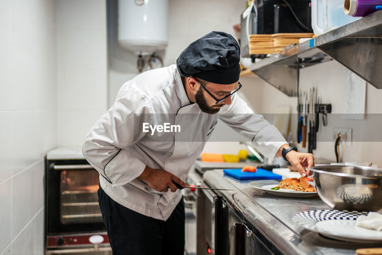 Concentrated male cook in uniform standing at metal counter in kitchen of cafe and preparing delicious dish with salmon fish