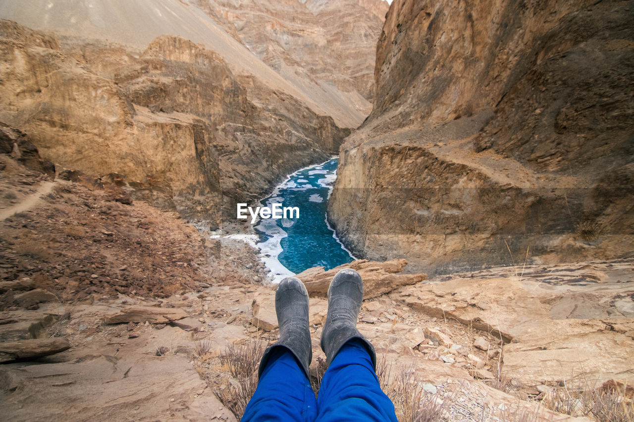 View of human legs wearing rubber boots and stream flowing through rocky mountain