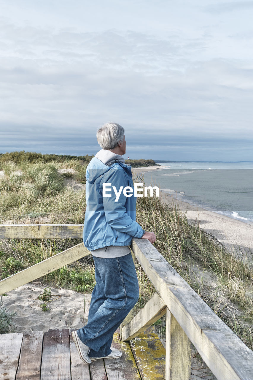 Senior woman with short gray hair in blue jeans and windbreaker standing on wooden platform by sea.