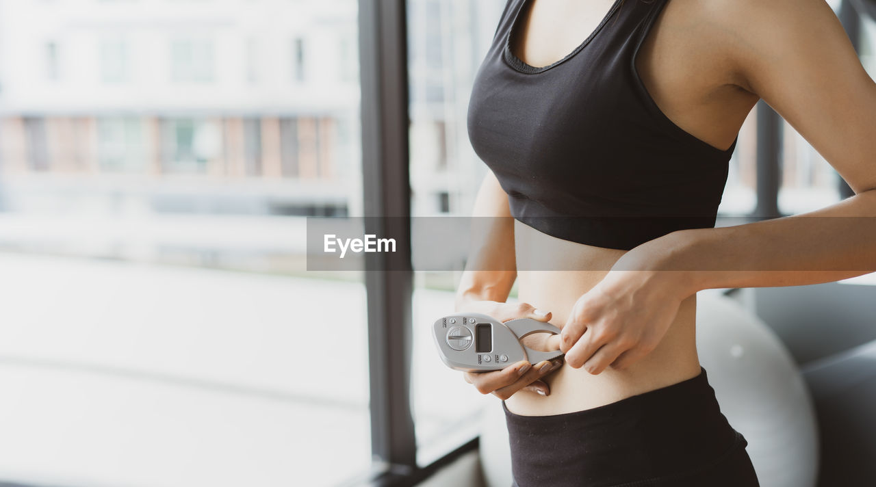 MIDSECTION OF WOMAN HOLDING CAMERA WHILE STANDING AGAINST WHITE WALL