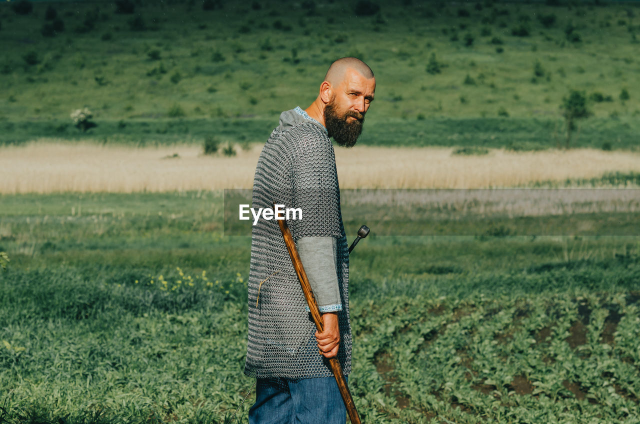 Bald bearded man in metal chain mail over linen shirt stands in middle of field, holding sword.