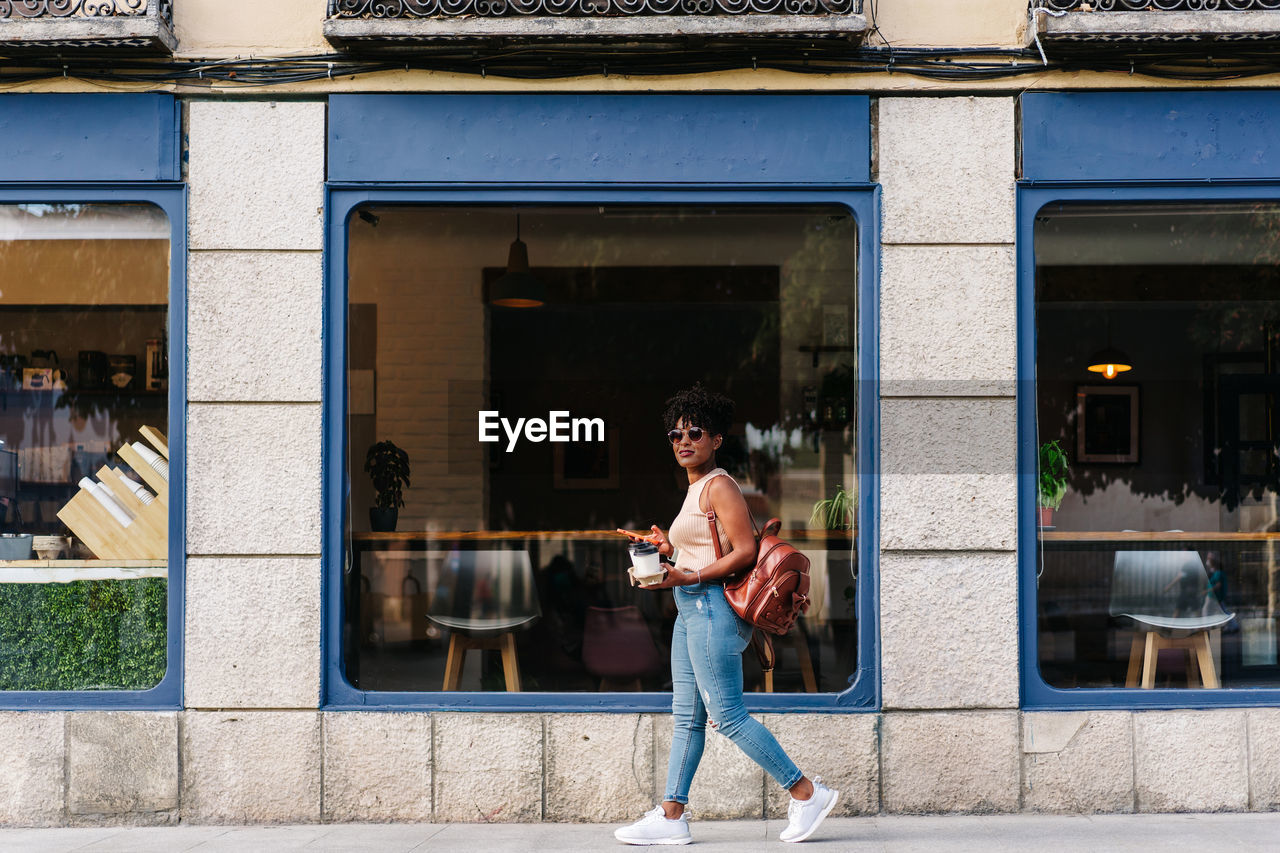 Full body of trendy young ethnic female millennial with dark afro hair in stylish outfit and backpack using mobile phone while walking on city street with cups of takeaway coffee