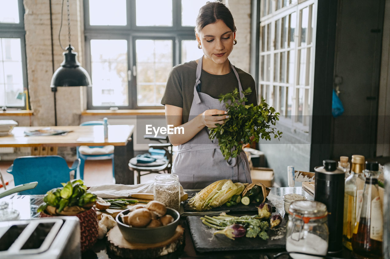 Female chef wearing apron doing quality check of cilantro standing in studio kitchen