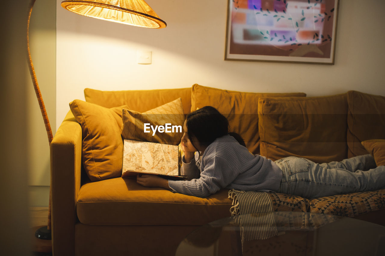 Side view of girl reading book under illuminated lamp while lying on sofa at home