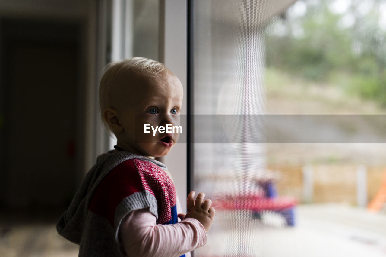 Girl with mouth open looking away while standing by window at home