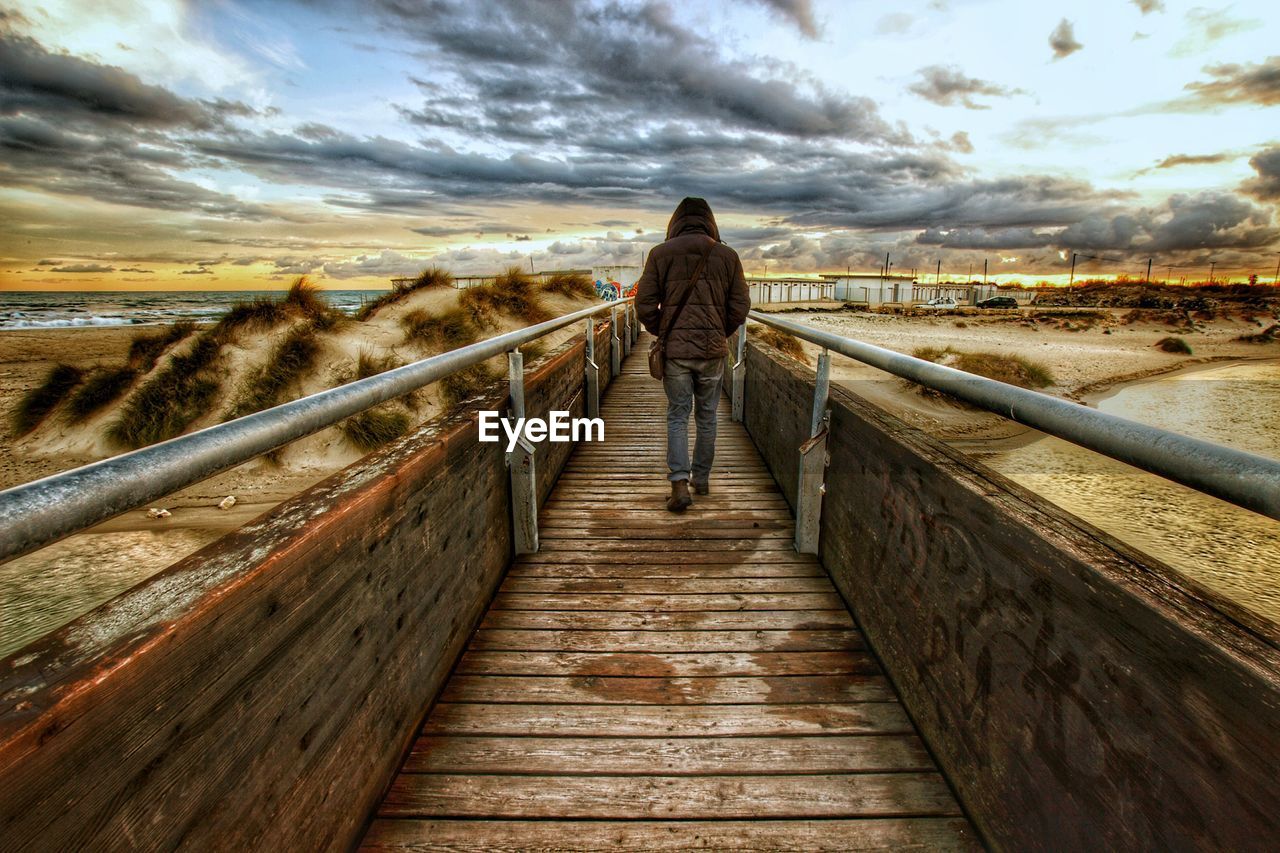 REAR VIEW OF WOMAN WALKING ON WOODEN RAILING AGAINST SKY