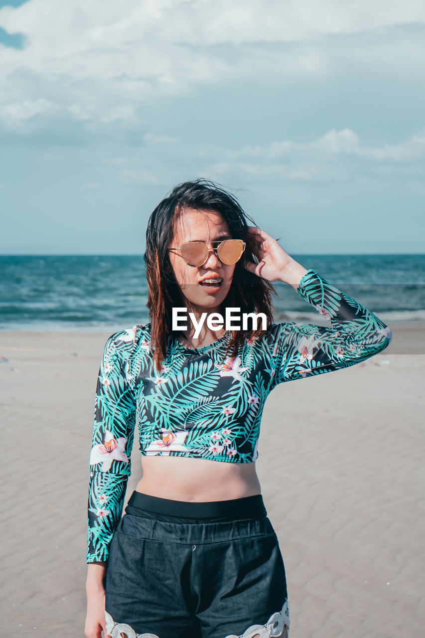 Young woman wearing sunglasses while standing at beach against sky during sunny day