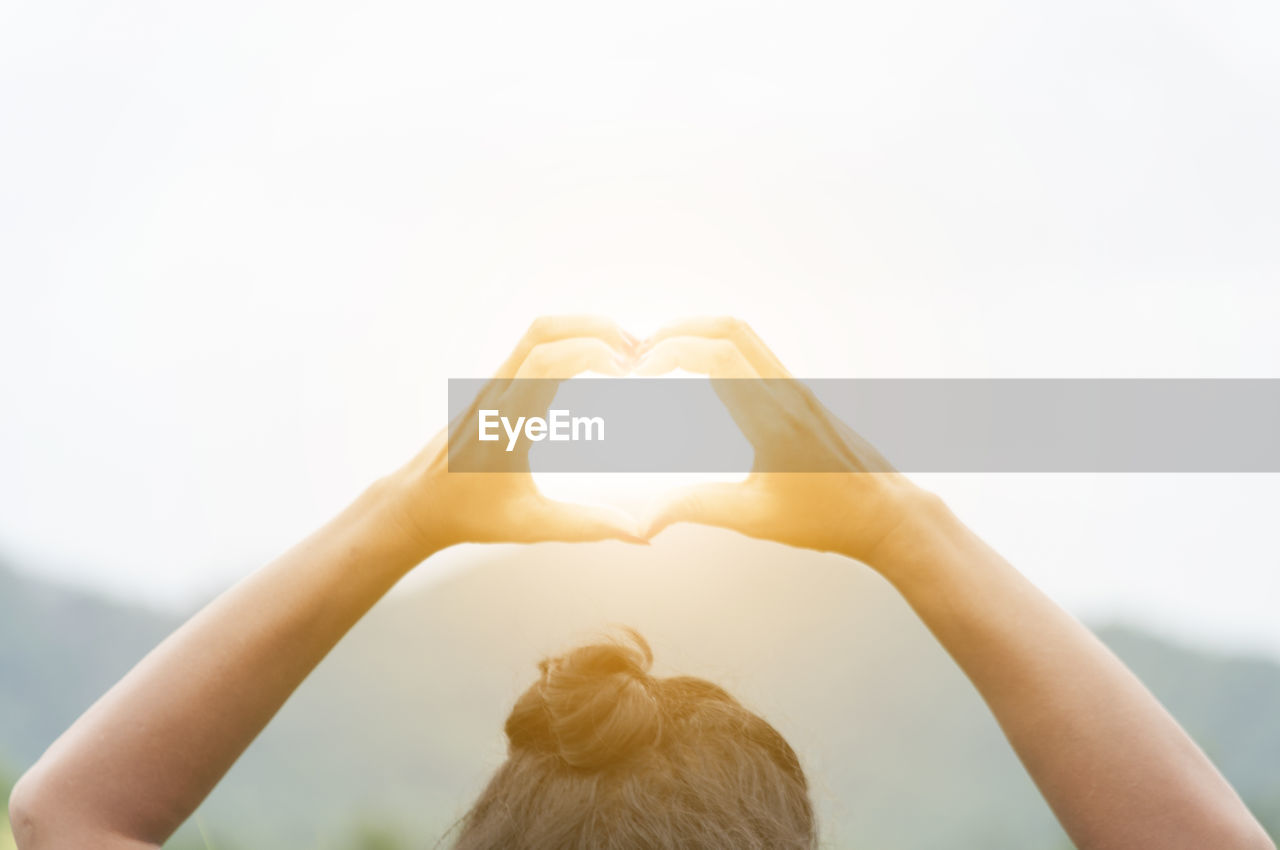 Cropped image of woman making heart shape against bright sun