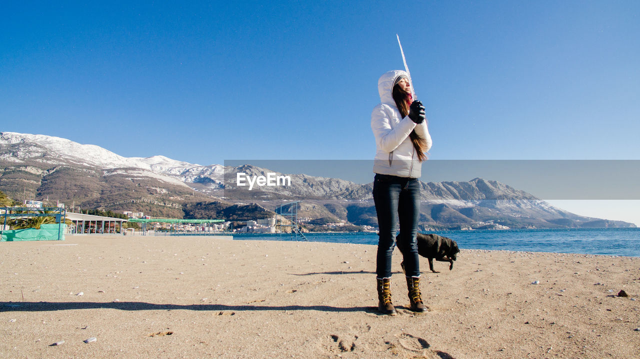 Woman holding icicle while standing with dog at beach against sky