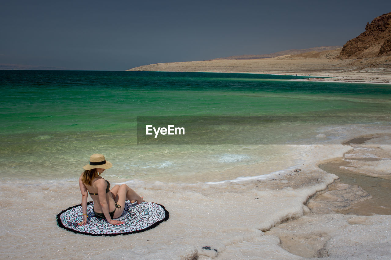 YOUNG WOMAN SITTING ON BEACH AGAINST SEA