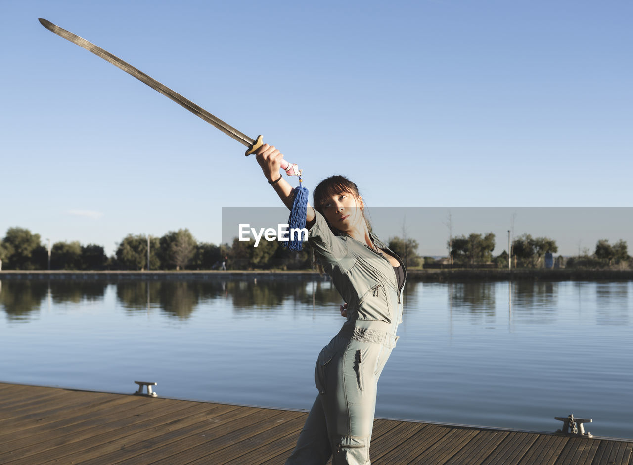 Young woman doing martial arts with sword in park by lake on sunny day