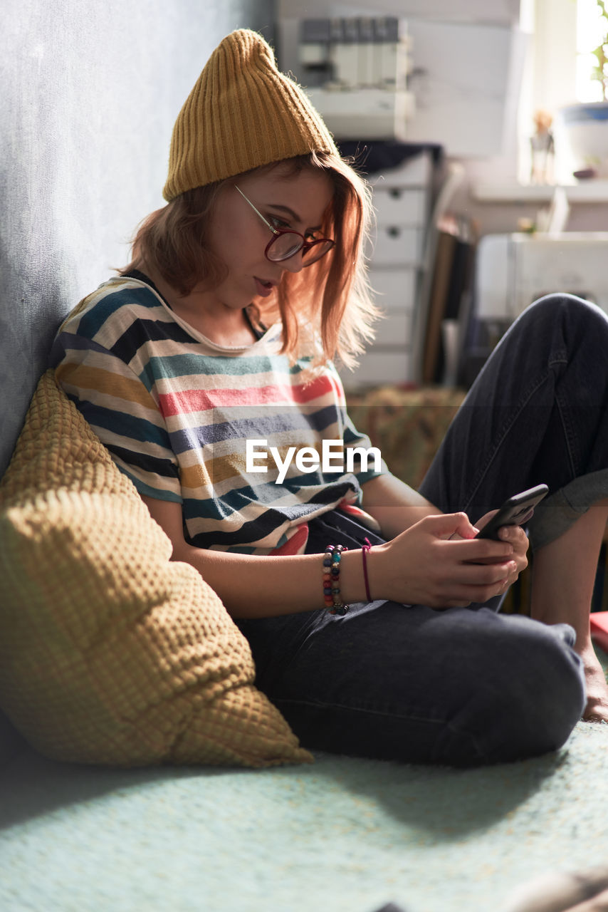 Side view of female millennial in trendy wear sitting on sofa at home and chatting with friends on social media via mobile phone