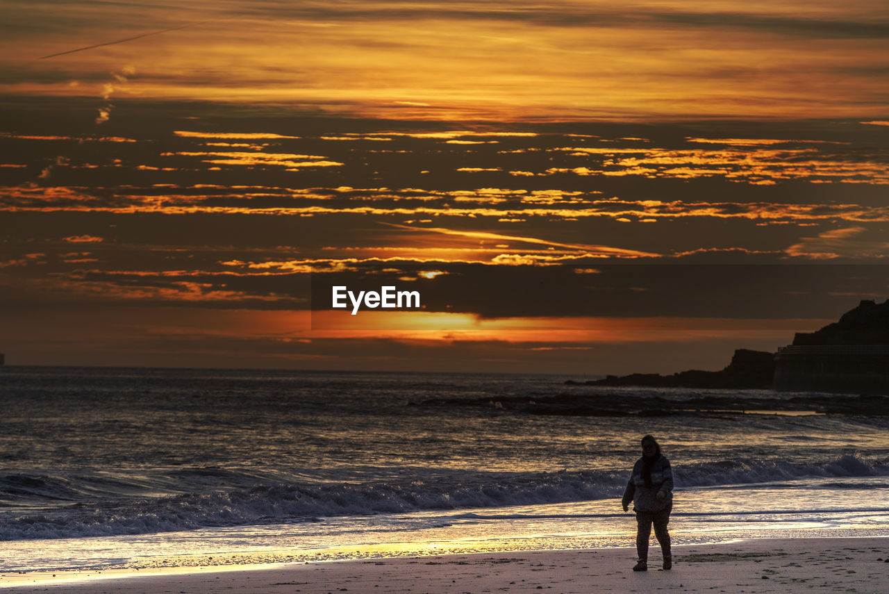 FULL LENGTH OF WOMAN STANDING ON BEACH DURING SUNSET