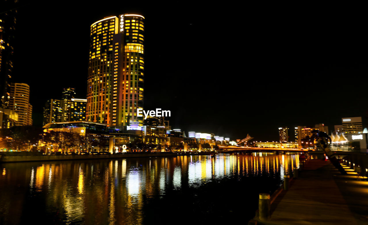 ILLUMINATED CITY BUILDINGS BY RIVER AGAINST SKY AT NIGHT