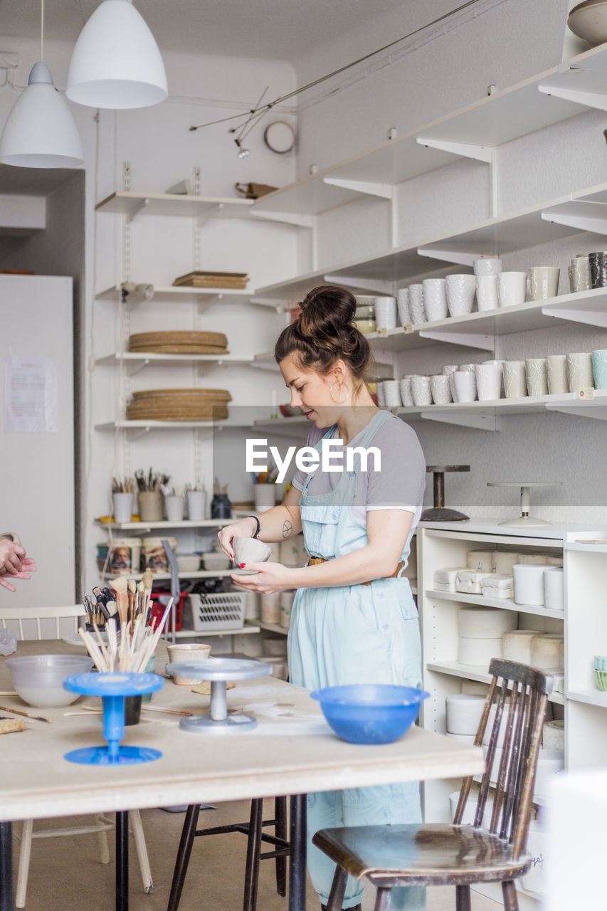 Young female potter molding shape from clay while standing by workbench at store