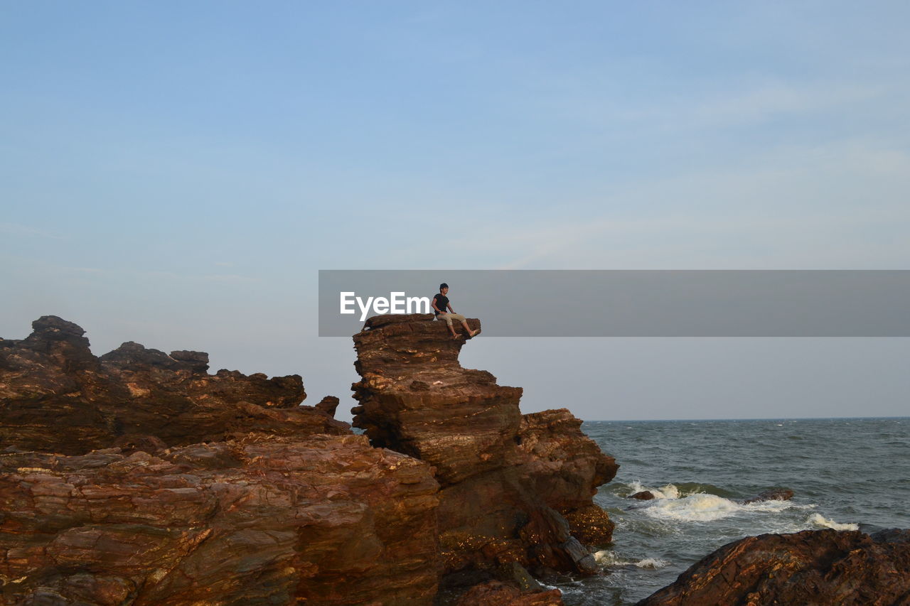 Low angle view of man sitting on cliff by sea against sky