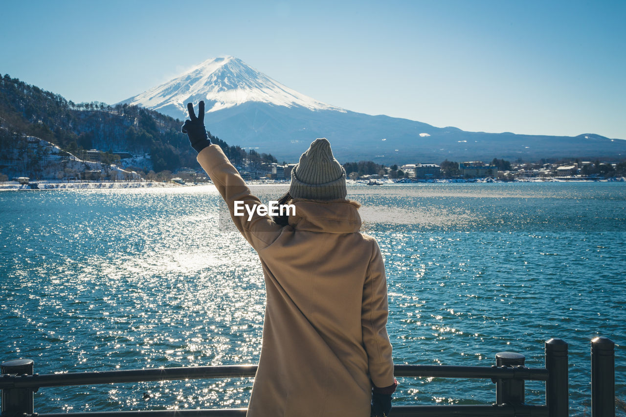 Rear view of woman gesturing peace sign while standing against lake in winter