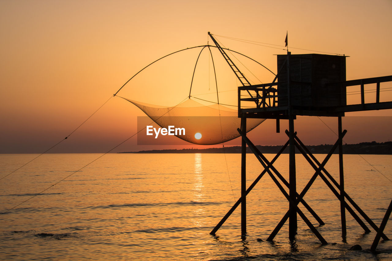 SILHOUETTE FISHING NET ON SEA AGAINST ORANGE SKY