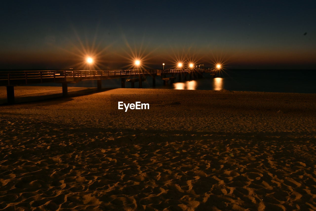 VIEW OF ILLUMINATED STREET AT BEACH AGAINST SKY AT NIGHT