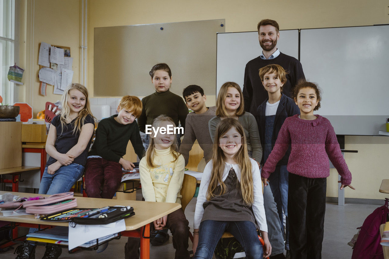 Portrait of smiling teacher and girls and boys against whiteboard in classroom