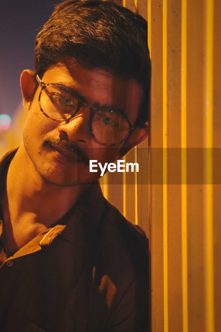 PORTRAIT OF YOUNG MAN WITH EYEGLASSES STANDING IN BACKGROUND