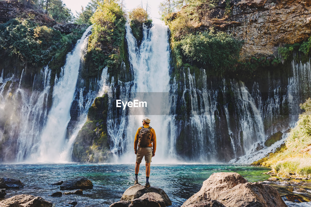 Man in a hat and yellow jacket looking on the beautiful waterfall, view from the back