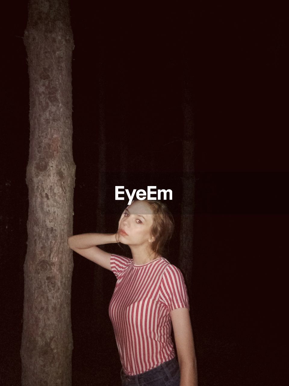 Portrait of young woman standing by tree trunk in forest at night