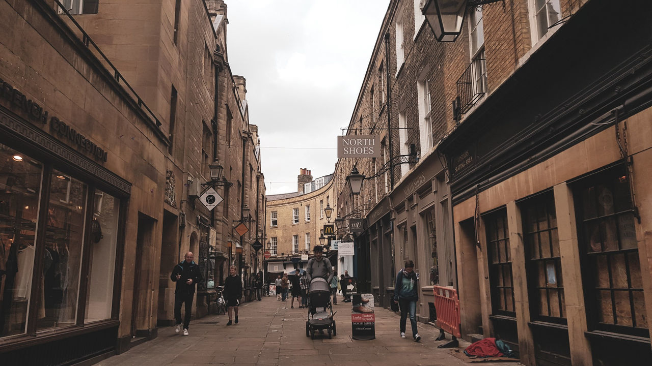 PEOPLE WALKING ON STREET AMIDST BUILDINGS IN CITY AGAINST SKY