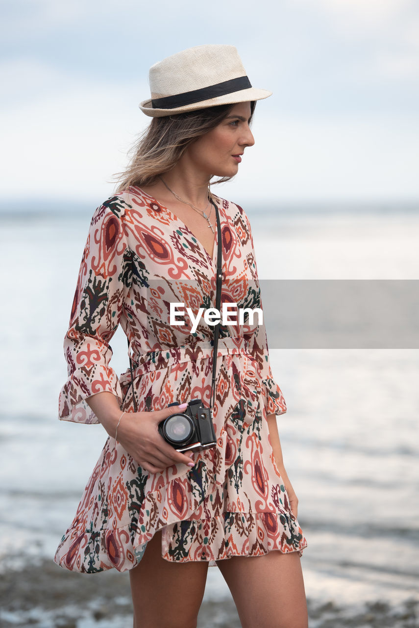 Fashionable caucasian woman holding camera and photographing scenery at the beach. 