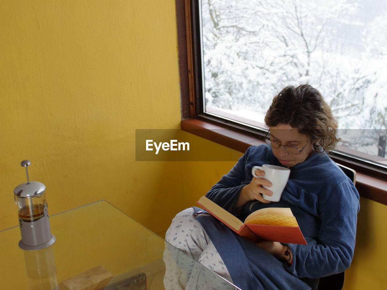 A caucasian woman in her pajamas drinks a cup of tea in front of the window as it snows outside